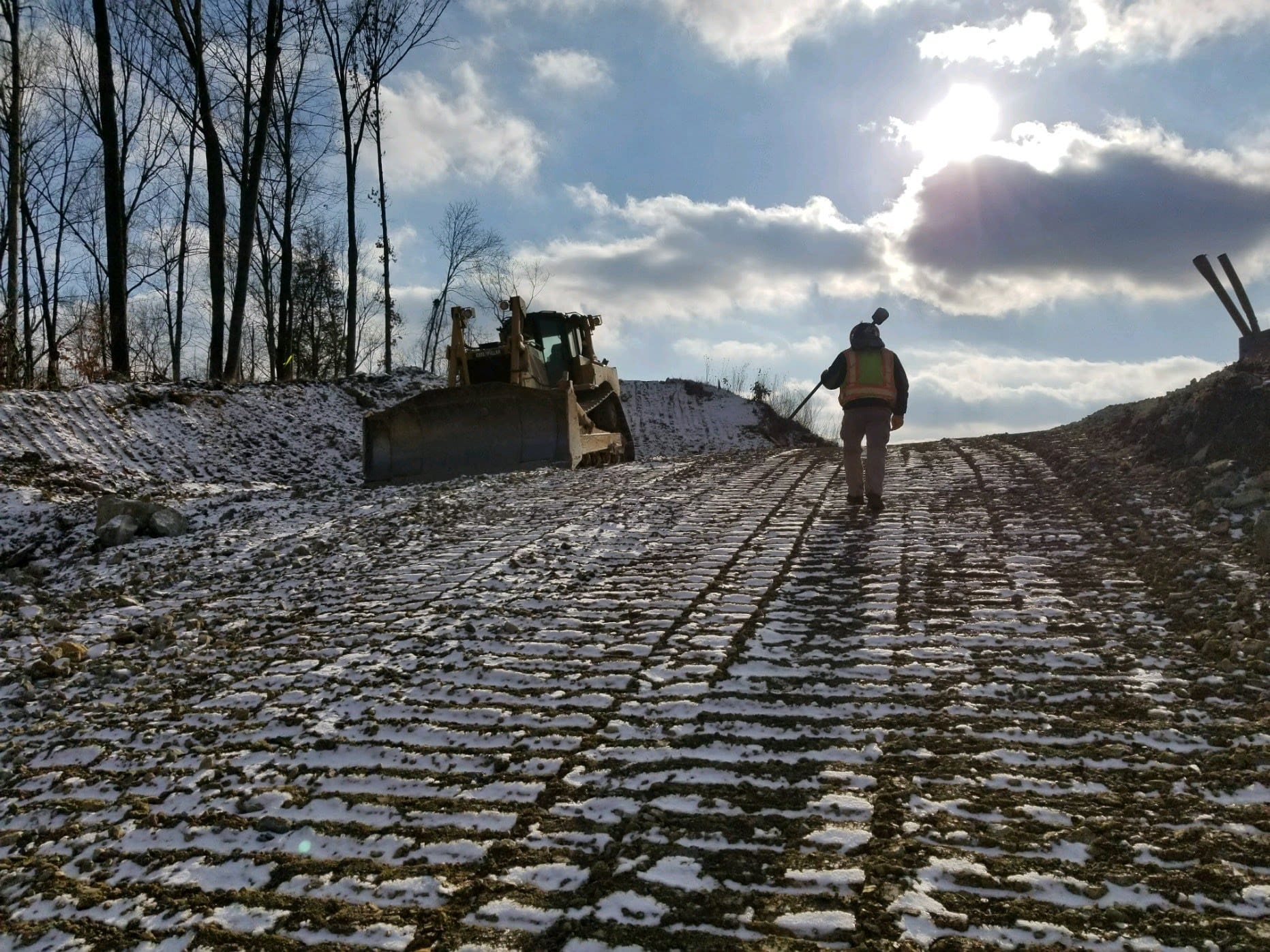 Surveyor walks up a snowy hill with a bulldozer in the background